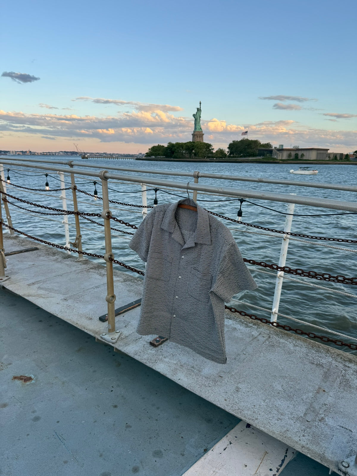 NYC Shirt in blue and white gingham seersucker on a hanger at the rail, Statue of Liberty behind
